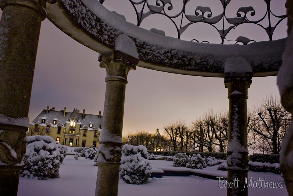 Verschneite Gartenarchitektur mit Rundbogen im Vordergrund, dahinter das Oheka Castle in der Abenddämmerung