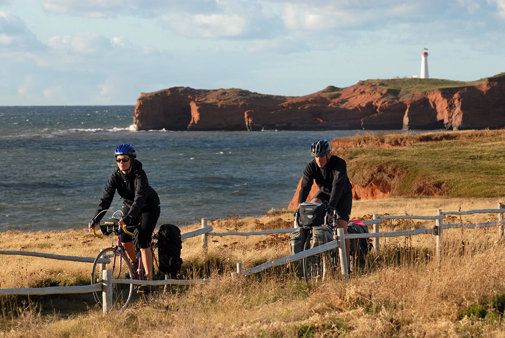 Radfahrer auf einem Küstenweg bei Wind und Wellen auf den Îles de la Madeleine mit roter Steilküste und Leuchtturm im Hintergrund.