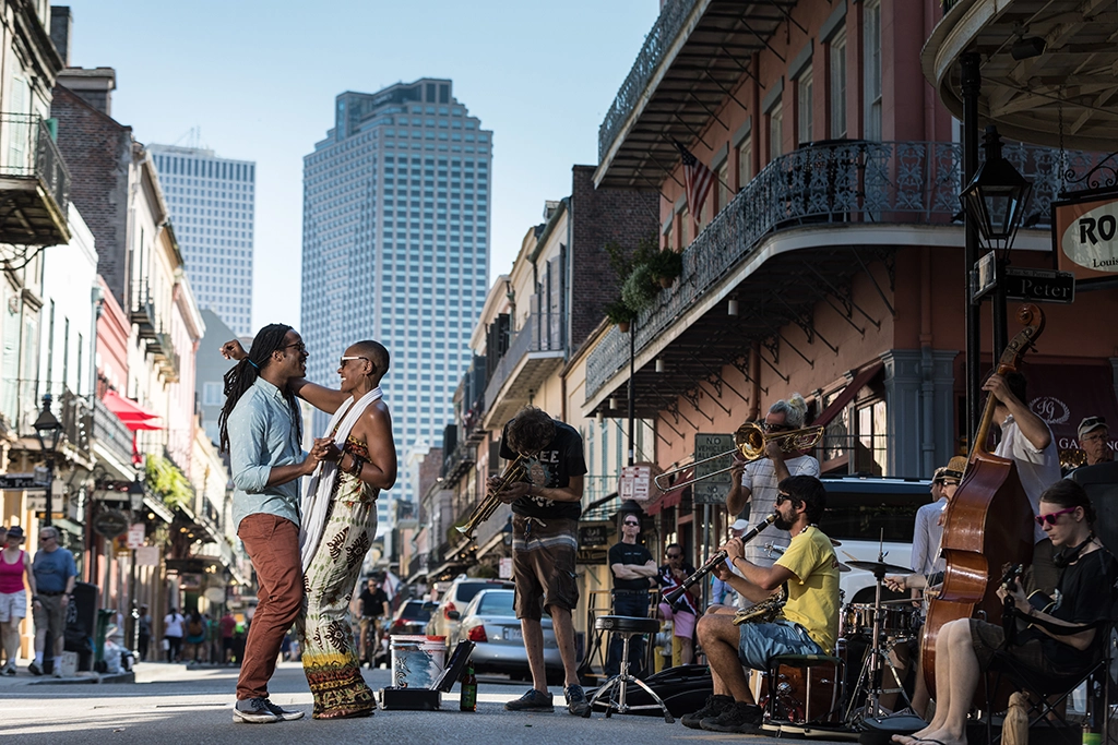 Ein Paar tanzt zu Livemusik einer Brass Band in der Royal Street in New Orleans, während die Sonne zwischen historischen Balkonen und modernen Hochhäusern scheint.