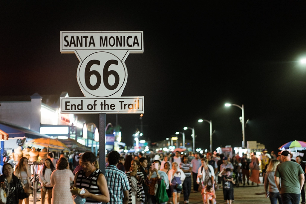 Das berühmte „End of the Trail“-Schild der Route 66 in Santa Monica leuchtet nachts über einer belebten Promenade.
