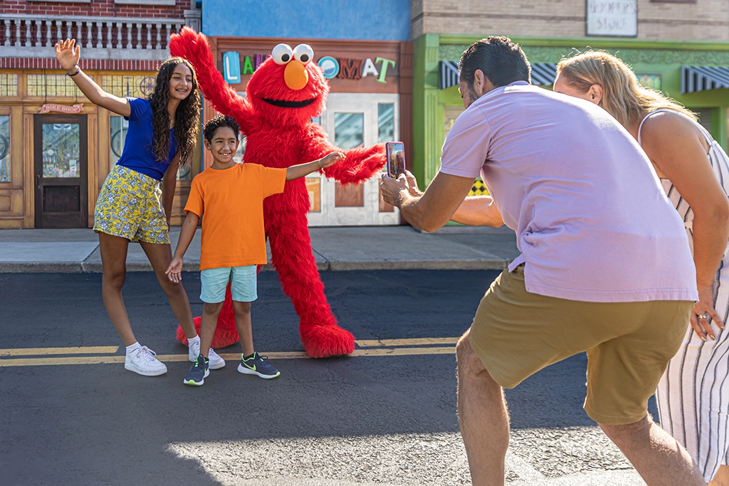 Zwei Kinder posieren für ein Foto mit Elmo, während Eltern sie lachend fotografieren, im Sesame Place Themenpark.