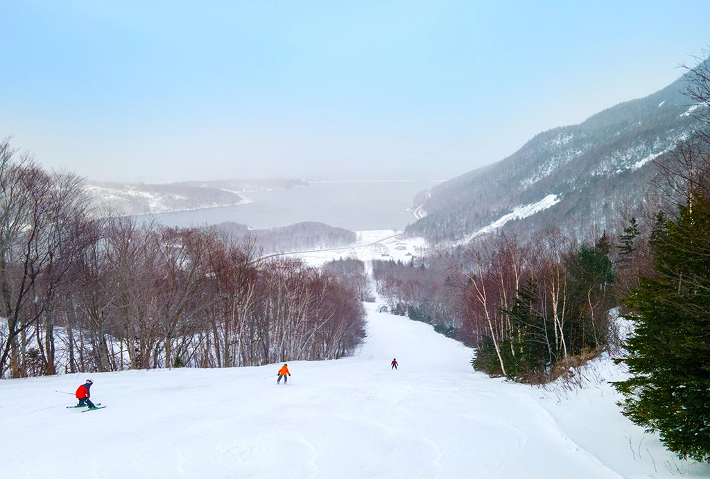 Skifahrer auf einer breiten Abfahrt mit Blick über den verschneiten Cape Smokey und den Atlantik in Nova Scotia.