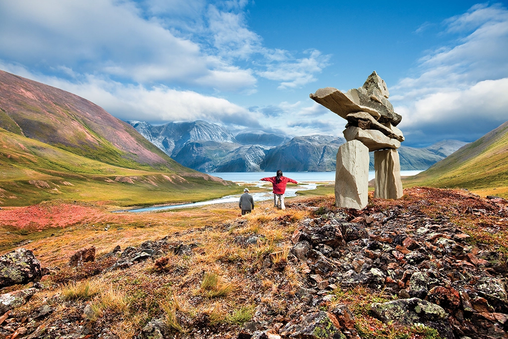 Zwei Wanderer stehen neben einem traditionellen Inukshuk im Torngat Mountains National Park in Labrador, mit Blick auf dramatische Berglandschaften und einen Fjord.