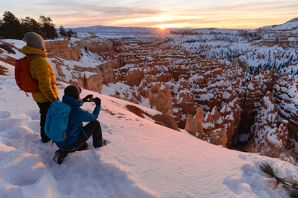Zwei Winterwanderer genießen den Sonnenuntergang über den verschneiten Felsformationen des Bryce Canyon Nationalparks.