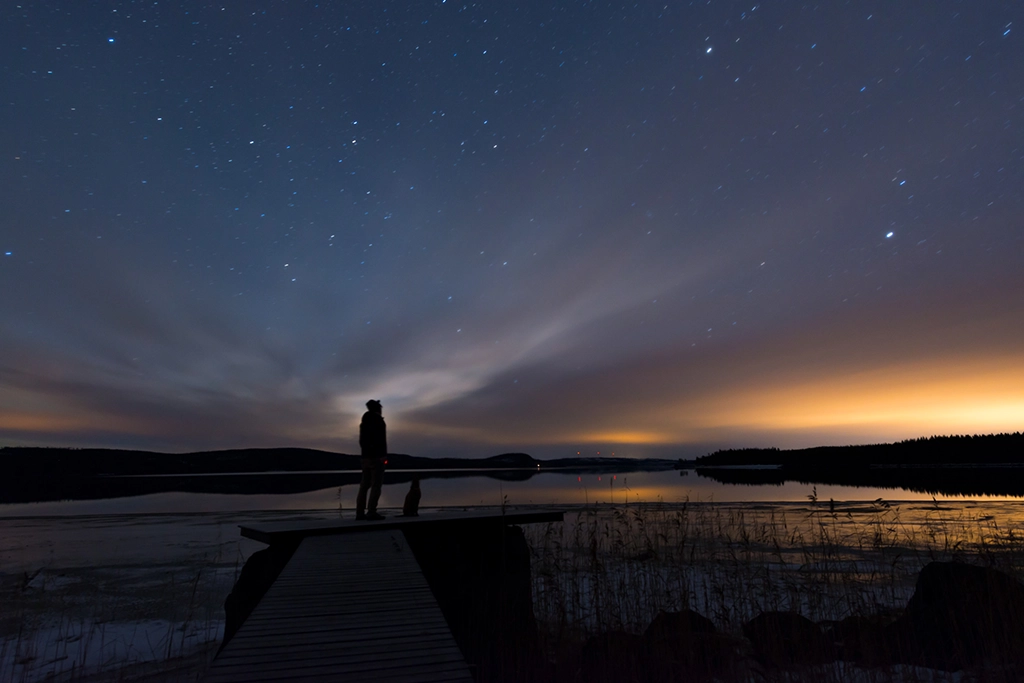 Person mit Hund auf einem Steg betrachtet den Sternenhimmel über einem ruhigen Wintersee.