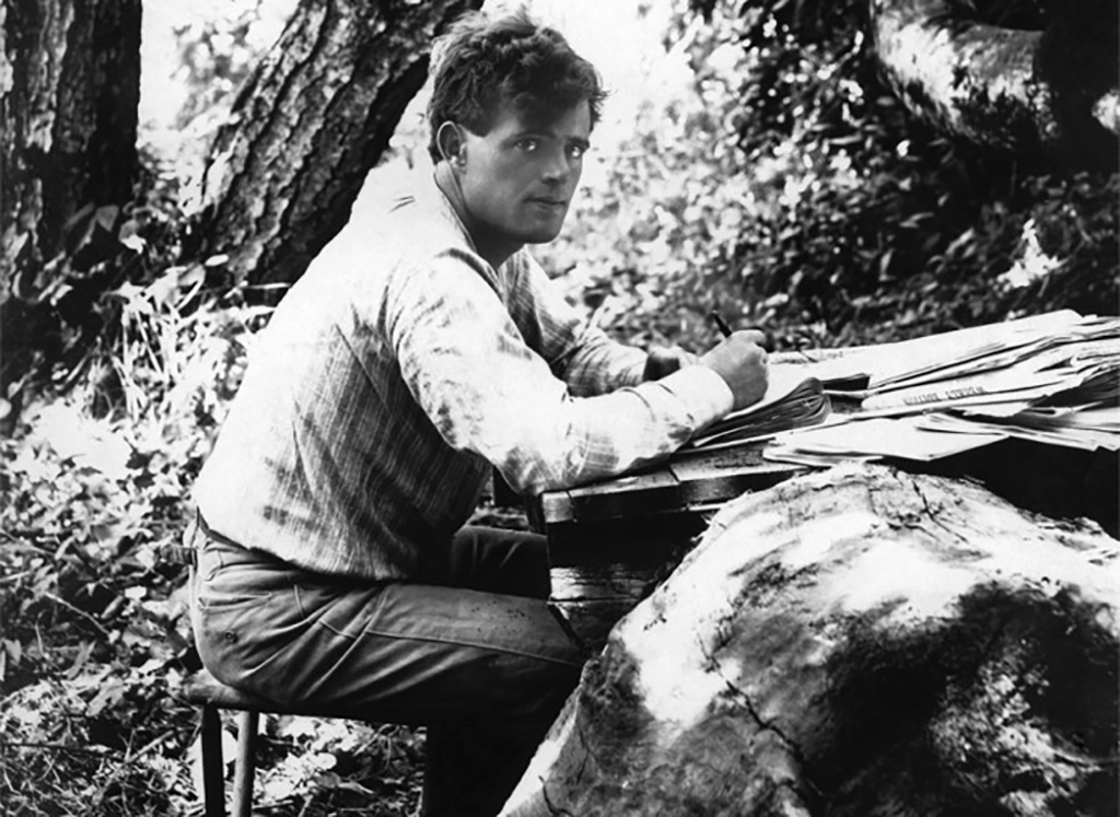 Jack London sitzt im Freien an einem Holztisch und schreibt, umgeben von Papierstapeln.