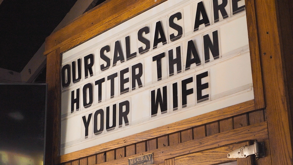 Leuchttafel mit Aufschrift „Our salsas are hotter than your wife“ in Holzrahmen