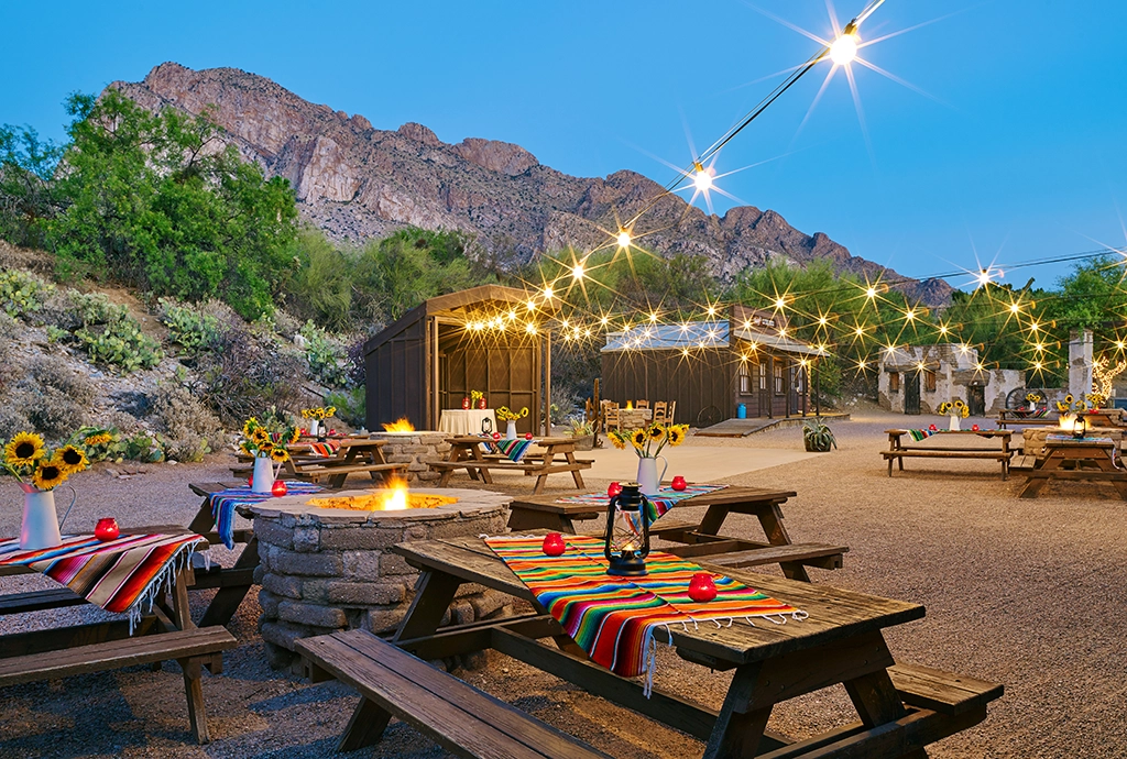 Lichterketten, Holztische mit bunten Decken und Lagerfeuerstelle in Outdoor-Atmosphäre am Fuße der Santa Catalina Mountains.
