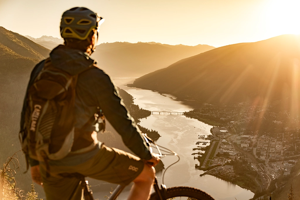 Mountainbiker blickt von einem Trail auf Nelson und den Kootenay Lake im Abendlicht.