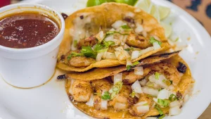 Zwei Tacos mit Garnelen, Zwiebeln und Koriander neben einem Becher Consomé