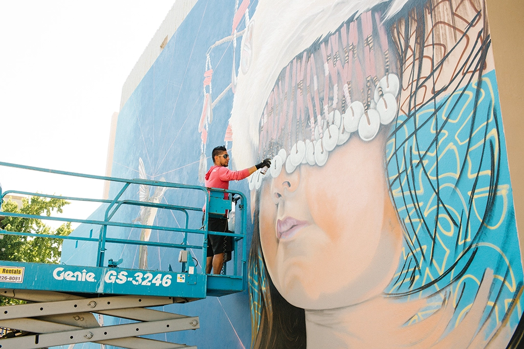Künstler malt ein großes Wandbild mit indigener Darstellung an einer Gebäudefassade.