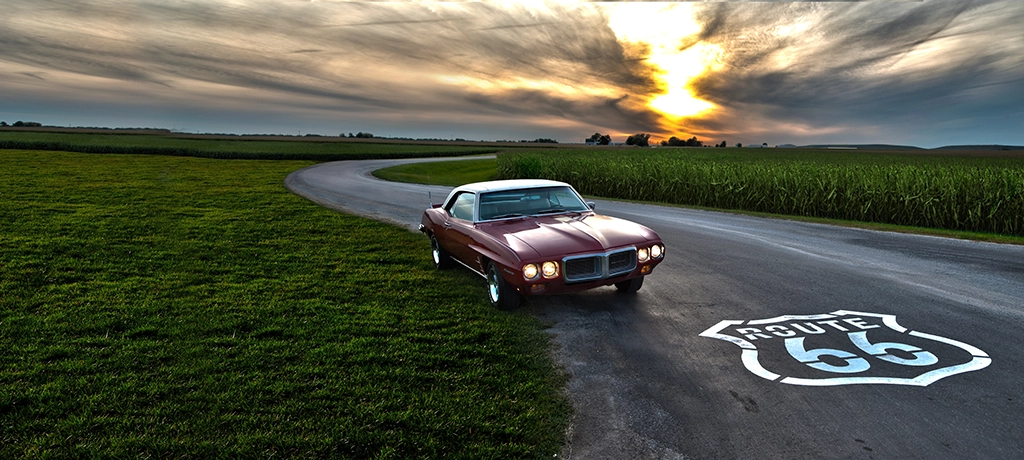 Klassischer Muscle Car auf der Route 66 im Sonnenuntergang neben Maisfeldern in Illinois.