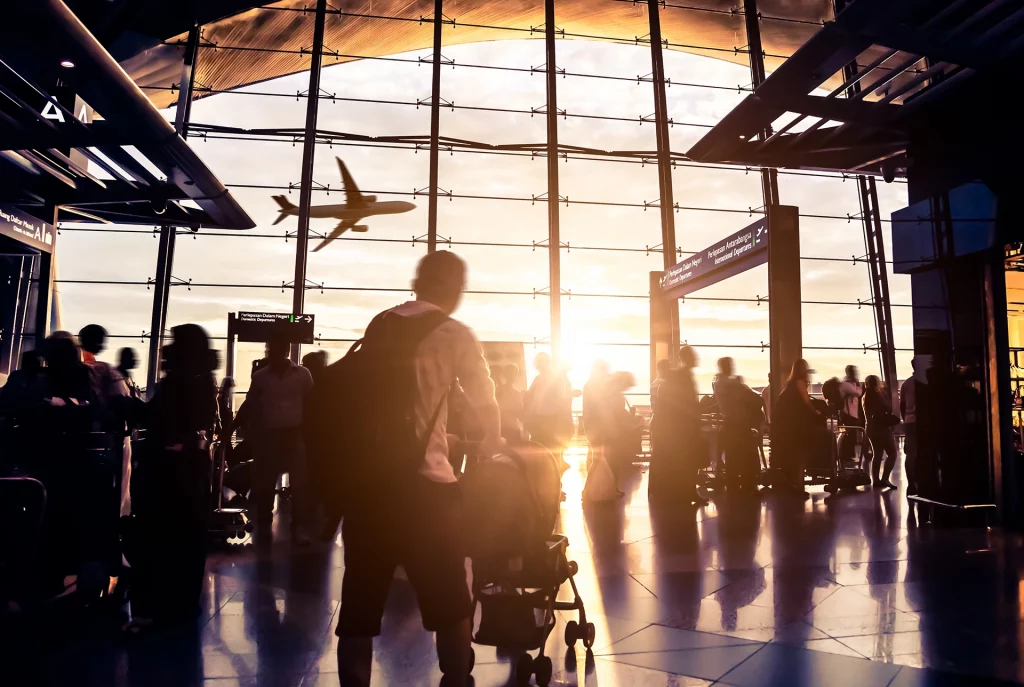 Reisende in einem internationalen Flughafen-Terminal bei Sonnenuntergang mit startendem Flugzeug.