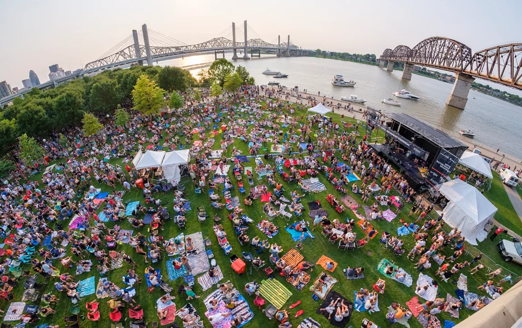 Besucher sitzen auf Decken im Waterfront Park bei einem Konzert am Ohio River.