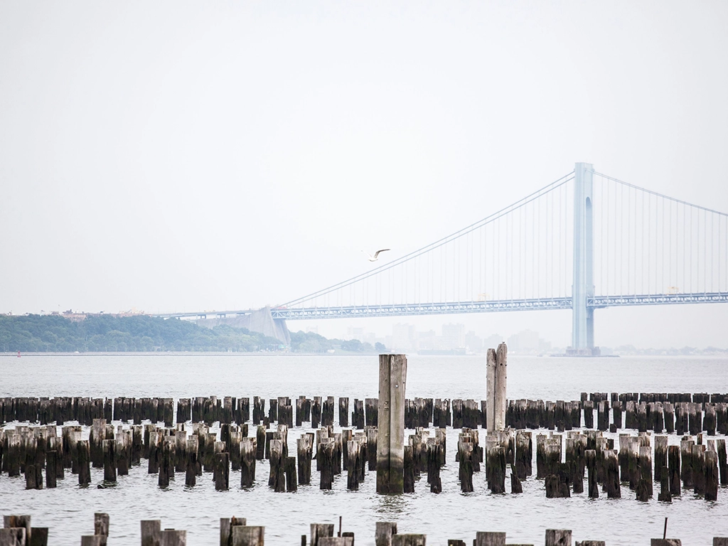 Blick von der Küste Staten Islands über alte Holzpfähle im Wasser auf die Verrazzano-Narrows Bridge zwischen Staten Island und Brooklyn.