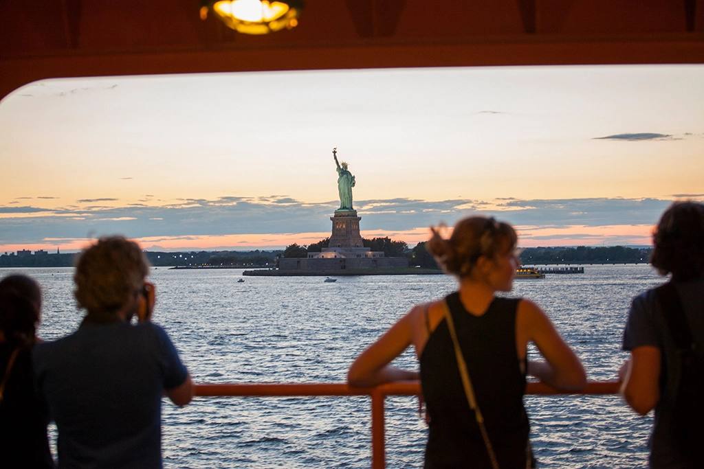 Passagiere auf der Staten Island Ferry blicken bei Sonnenuntergang über den New Yorker Hafen auf die Freiheitsstatue.