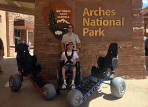 Zwei spezielle Geländerollstühle von Extreme Motus vor dem Besucherzentrum des Arches Nationalparks in Utah, daneben ein Rollstuhlfahrer mit Begleitperson.