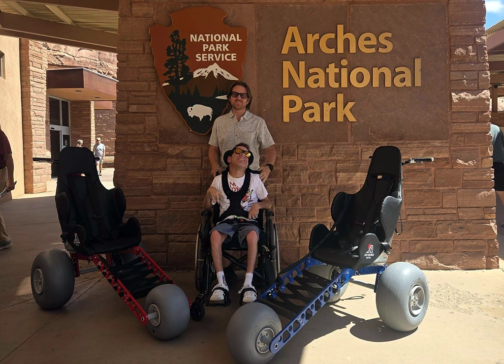 Zwei spezielle Geländerollstühle von Extreme Motus vor dem Besucherzentrum des Arches Nationalparks in Utah, daneben ein Rollstuhlfahrer mit Begleitperson.