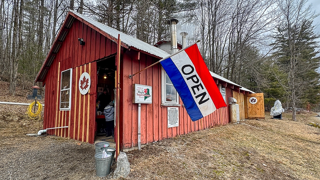 Rotes Sugarhouse der Cole Farm in Dayton, Maine, mit geöffneter Tür, Ahornblatt-Schildern und großer OPEN-Flagge