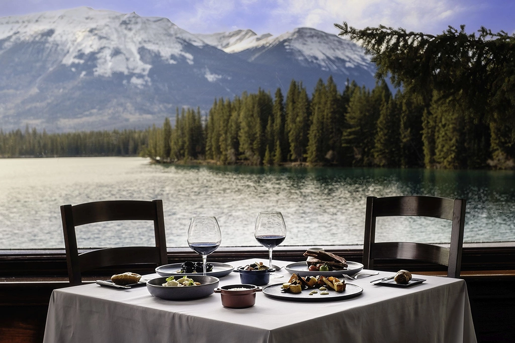 Gedeckter Restauranttisch mit Speisen und Rotwein in der Orso Trattoria der Fairmont Jasper Park Lodge mit Blick auf den Lac Beauvert und die Rocky Mountains