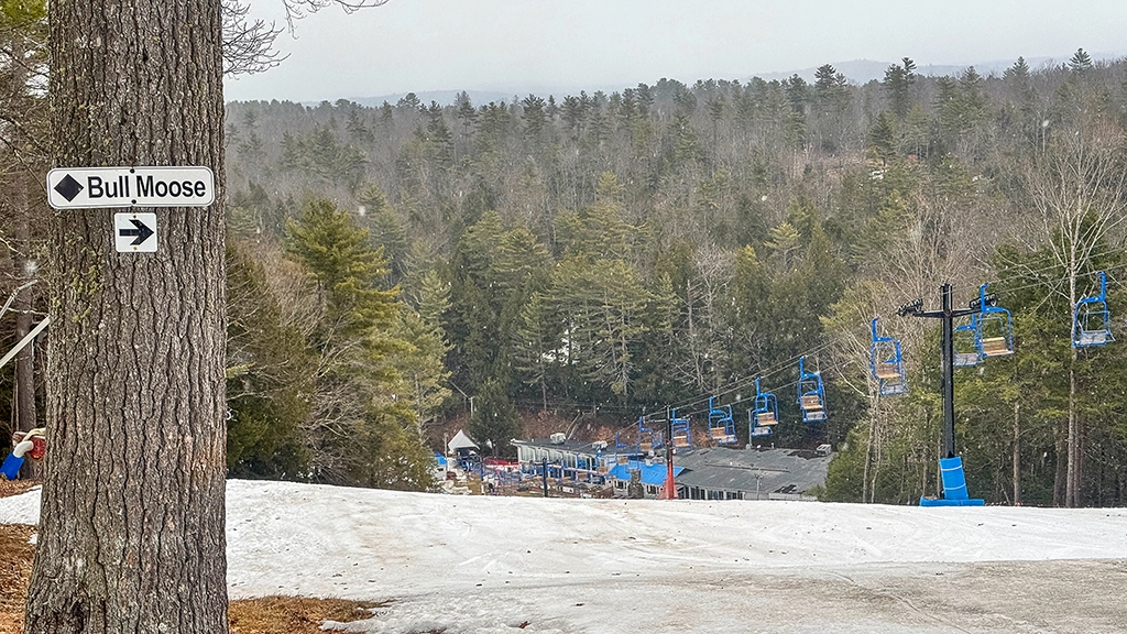 Blick von der Skipiste Bull Moose im Skigebiet Lost Valley bei Auburn in Maine auf blaue Sessellifte, die Talstation und bewaldete Hügel.