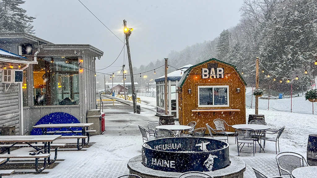 Starker Schneefall im Skigebiet Lost Valley in Maine mit kleiner Holzbar, Sitzgelegenheiten im Freien und beleuchteten Gebäuden.