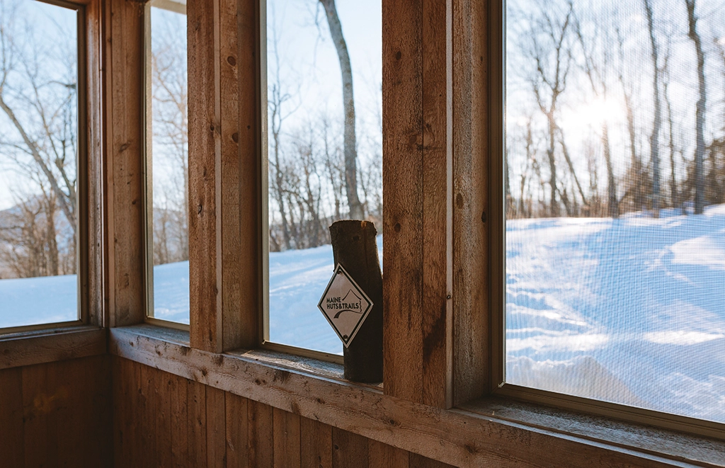 Blick durch die Holzfenster einer Hütte von Maine Huts & Trails auf eine verschneite Waldlandschaft in Maine