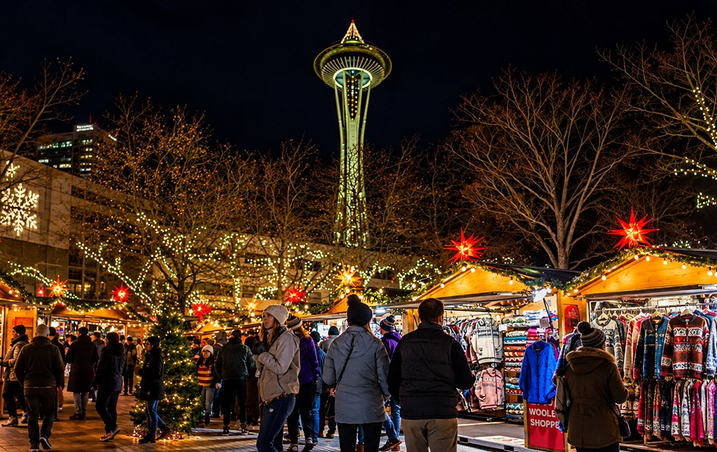 Spektakuläre Luftaufnahme der Space Needle in Seattle während der Weihnachtszeit. Die Spitze des berühmten Aussichtsturms ist mit zahlreichen Lichterketten festlich geschmückt. Dahinter erstreckt sich die Skyline von Downtown Seattle mit ihren beleuchteten Hochhäusern. Am Horizont erhebt sich der schneebedeckte Mount Rainier, während rechts die Elliott Bay, das Seattle Great Wheel und die farbig beleuchteten Bögen des Lumen Field zu erkennen sind. Das Foto verbindet die weihnachtliche Atmosphäre der Stadt mit ihrer eindrucksvollen Lage zwischen Wasser und Bergen.