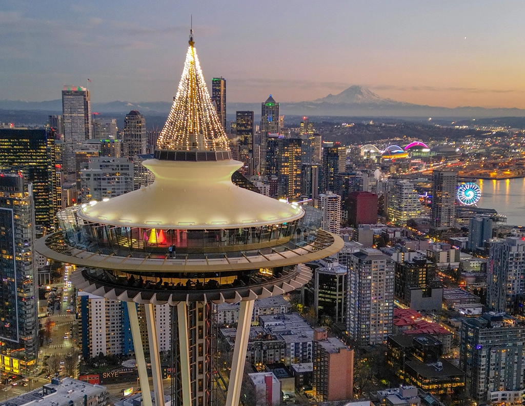 Weihnachtlich beleuchtete Space Needle über der Skyline von Seattle bei Sonnenuntergang, mit Mount Rainier und der Elliott Bay im Hintergrund.